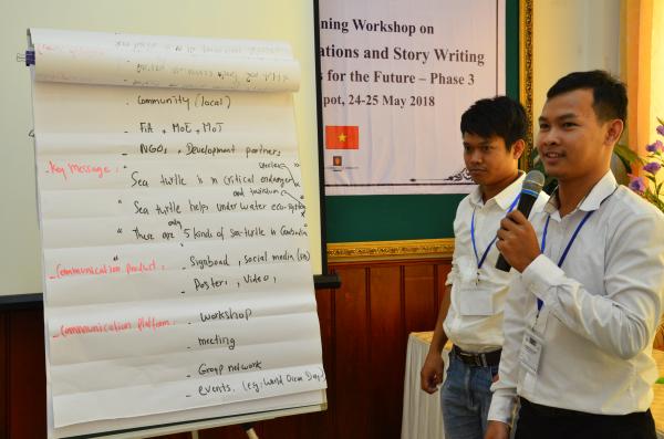 Two men stand near a pad of presentation paper. One speaks into a microphone.